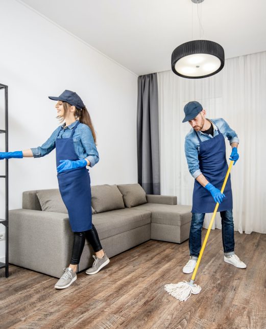 Professional cleaners in blue uniform washing floor and wiping dust from the furniture in the living room of the apartment. Cleaning service concept